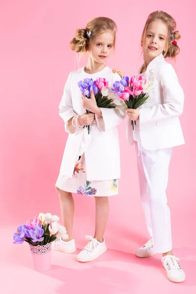 Gemelas en ropa ligera con ramos de flores de pie sobre un fondo rosa . — Foto de Stock