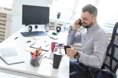 Ofiste genç bir adam üstünde belgili tanımlık telefon ve dökme kahve bir fincan içine konuşmak bir masada oturur.
