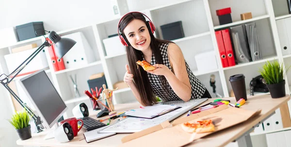 Uma jovem está perto de uma mesa, segura um pedaço de pizza na mão e mostra um sinal de classe. Antes que a rapariga em cima da mesa seja um quadro magnético. Na cabeça da menina usando fones de ouvido . — Fotografia de Stock