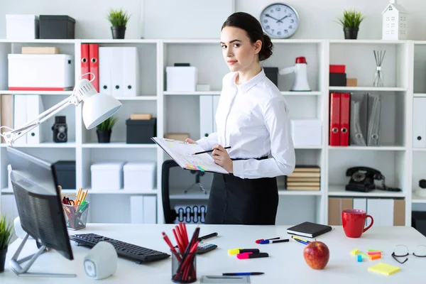 Una joven en la oficina está de pie cerca de la mesa y sostiene en sus manos una sábana para notas y un lápiz . — Foto de Stock