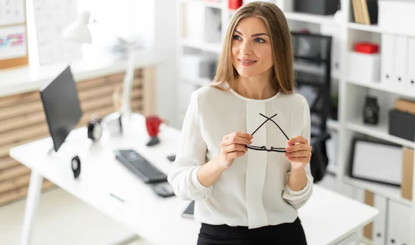 Seorang gadis muda berdiri bersandar di meja di kantor dan memegang kacamata di tangannya . — Stok Foto