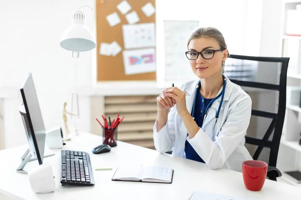 Ein junges Mädchen im weißen Gewand sitzt an den Tischen im Büro und hält einen Stift in der Hand. Ein Stethoskop hängt um ihren Hals. — Stockfoto