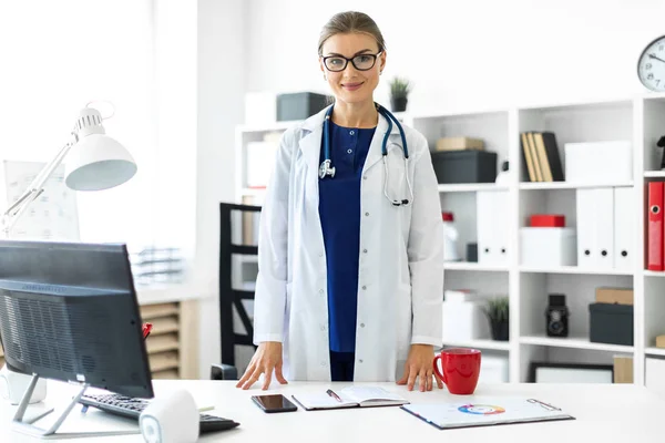 Una giovane ragazza con un cappotto bianco è in piedi vicino a un tavolo nel suo ufficio. Uno stetoscopio le pende intorno al collo . — Foto Stock