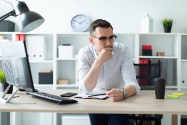 Gözlüklü genç bir adam ofiste bir tabloda yakınındaki duruyor ve bir kalem elinde tutuyor.