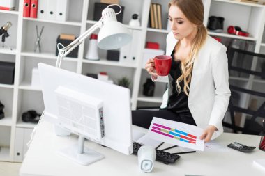 Genç kız ofisinde, masada kırmızı bir fincan elinde tutan ve bir bilgisayar ve belgeleri çalışma oturdu.