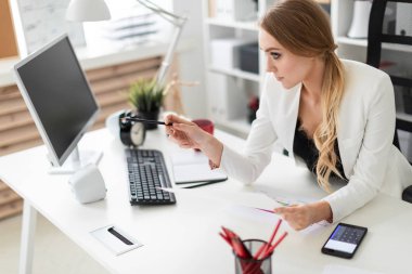 Genç bir kız ofiste, bilgisayar masasında oturuyor onun elinde kalem tutan ve belgeleriyle çalışma.