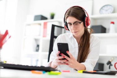 Genç bir kız kulaklık ofiste bir masada oturur ve bir telefon elinde tutan.