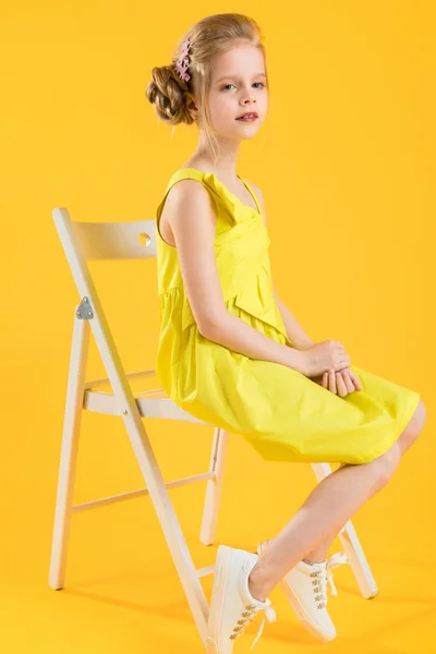 Una chica está sentada en una silla blanca sobre un fondo amarillo . — Foto de Stock