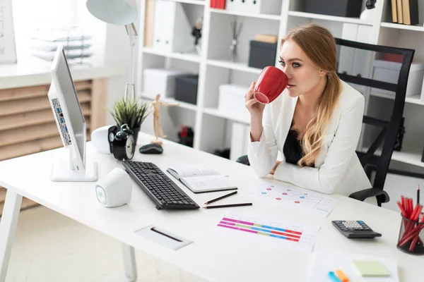 Mladá dívka sedí u počítače, recepce v kanceláři a má červený hrnek v ruce. Před dívku na stůl jsou diagramy. — Stock fotografie