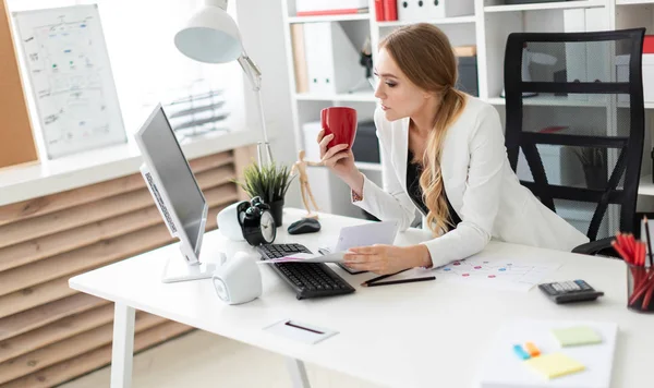 Una joven está sentada en el escritorio de la computadora en la oficina y sostiene una taza y documentos. Antes de que la chica en la mesa son diagramas . — Foto de Stock