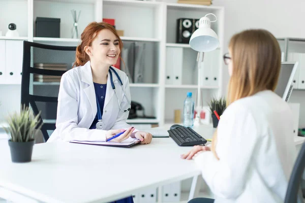 Uma linda menina em um manto branco está sentada na mesa no escritório e se comunicando com o interlocutor. A menina faz anotações na folha . — Fotografia de Stock