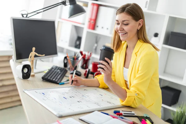 A young girl stands near a table and holds a glass of coffee and a marker. Before the girl on the table is a magnetic board — Stock Photo, Image