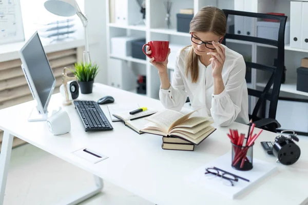 Una joven se sienta en una mesa en la oficina y sostiene una taza roja en su mano y lee un libro . — Foto de Stock