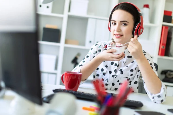 Belle jeune fille assise dans des écouteurs au bureau en mangeant du yaourt avec remplissage rouge . — Photo