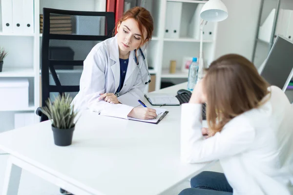 A beautiful young girl in a white robe is sitting at the desk in the office and communicating with the interlocutor. The girl makes notes in the sheet. — Stock Photo, Image