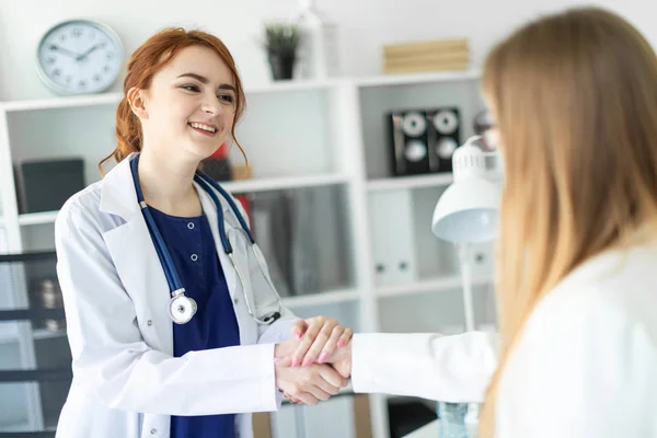 Uma menina bonita com um casaco branco está de pé perto da mesa no escritório e apertando as mãos com o paciente. O paciente agradece ao médico . — Fotografia de Stock