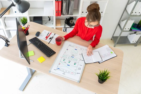 Una joven está sentada en una mesa de la oficina, sosteniendo una taza roja en la mano y mirando el bloc de notas. Un tablero magnético yace delante de la chica . —  Fotos de Stock
