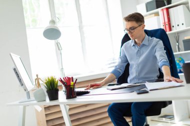 Genç bir adam bir bilgisayar masası ofisinde oturuyor ve bir kalem elinde tutuyor.