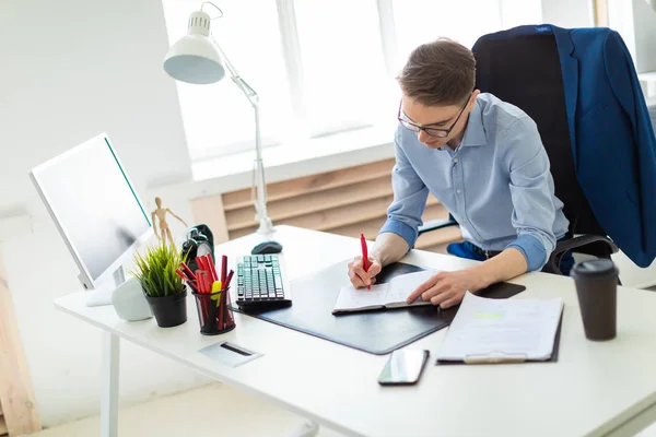 Un joven se sienta en la oficina en un escritorio de computadora y escribe en un cuaderno . — Foto de Stock