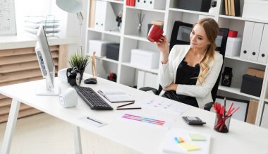 Genç bir kız ofiste bilgisayar masasında oturur ve kırmızı bir fincan elinde tutar. Kız masada daha önce diyagram olan.