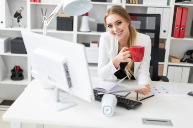 Genç bir kız ofiste bilgisayar masasında oturuyor ve bir fincan ve belgeleri tutuyor. Kız masada daha önce diyagram olan.