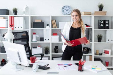 Masanın yanındaki ofiste duran genç bir kız elinde belgelerle bir dosya tutuyor..