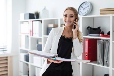 Genç bir kız ofis raf yanında duran, telefonda konuşurken ve belgeler ile bir klasör holding.