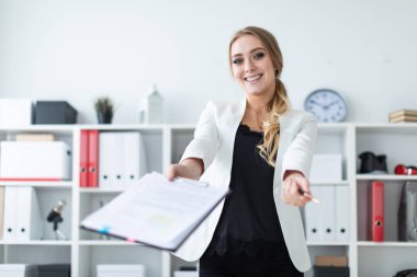Genç bir kız ofis raf yanında duruyor ve belgeleri ve kalem tutar.