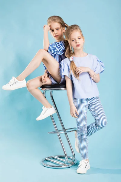 Mädchen-Zwillinge in hellblauer Kleidung posieren in der Nähe eines Barhockers auf blauem Hintergrund. — Stockfoto