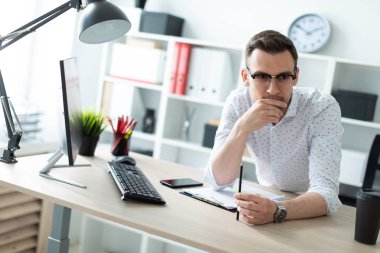 Gözlüklü genç bir adam ofiste bir tabloda yakınındaki duruyor ve bir kalem elinde tutuyor.