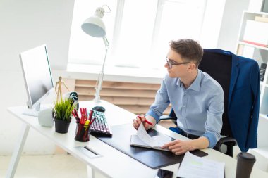 Genç bir adam bilgisayar masası ofisinde oturuyor, bir kalem elinde tutuyor ve ekrana görünüyor.