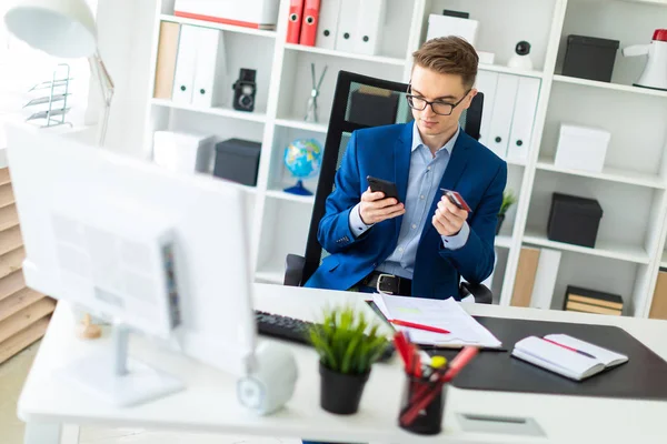 Молодой человек сидит за столом в офисе и держит банковскую карточку и телефон . — стоковое фото