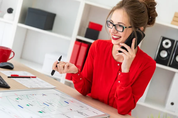 Una joven está de pie en una mesa en la oficina, sosteniendo un marcador negro en su mano y hablando por teléfono. La chica trabaja con una computadora, bloc de notas y una placa magnética . — Foto de Stock