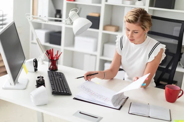 Молодая блондинка сидит за компьютерным столом в офисе, держит карандаш в руке и просматривает документы. . — стоковое фото