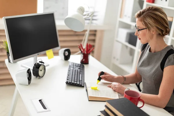 En ung flicka sitter vid ett bord på kontoret, hålla en gul markering i handen. Innan flickan ligger en öppen bok. — Stockfoto