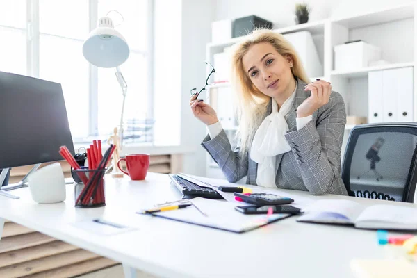 Una joven está sentada en una mesa en la oficina. La chica lleva gafas en la mano. . — Foto de Stock