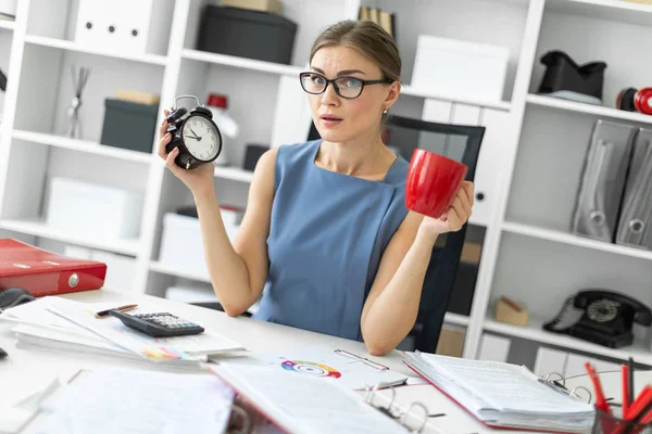 Uma jovem está sentada em uma mesa em seu escritório, segurando um despertador e um copo vermelho . — Fotografia de Stock