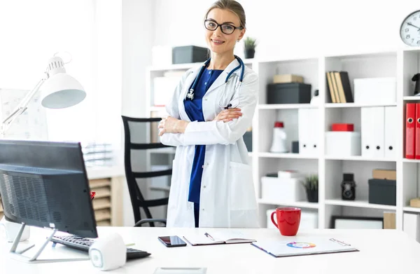 Una giovane ragazza con un cappotto bianco è in piedi vicino a un tavolo nel suo ufficio. Uno stetoscopio le pende intorno al collo . — Foto Stock