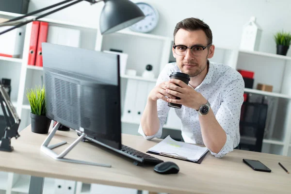 Молодой человек в очках стоит возле стола в офисе, держа стакан кофе . — стоковое фото