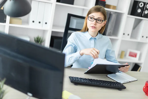 En ung pige med briller sidder ved et bord på kontoret, holder en blyant i hånden og arbejder med dokumenter. . - Stock-foto