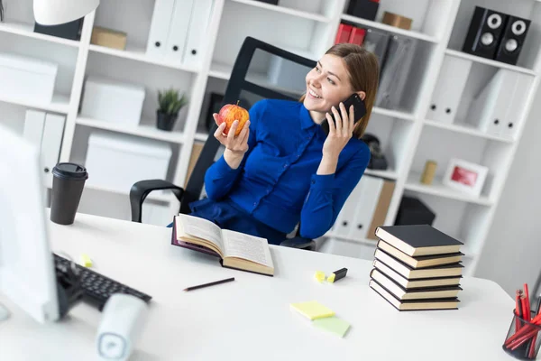 Молодая девушка сидит за компьютерным столом, держа в руке яблоко и разговаривая по телефону. Прежде чем девушка лежит открытая книга . — стоковое фото