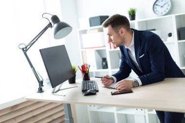 Genç bir adam ofiste bir tabloda yakınındaki duruyor, bir kalem elinde tutuyor ve belgeler ve bir bilgisayar ile çalışır.