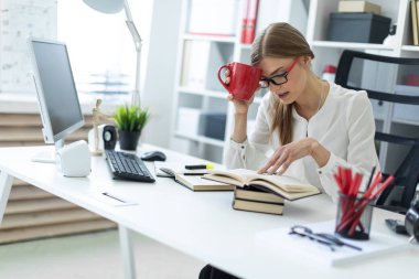 Genç bir kız ofiste bir masada oturur ve kırmızı bir fincan elinde tutan ve bir kitap okur.