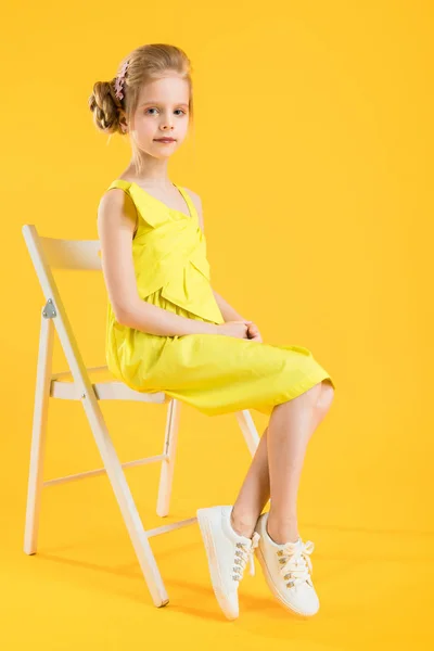 Una chica está sentada en una silla blanca sobre un fondo amarillo . — Foto de Stock