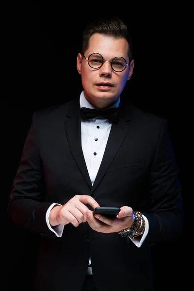 Porträt eines selbstbewussten gutaussehenden eleganten Geschäftsmannes mit Fliege in Brille und Telefon in der Hand auf schwarzem Hintergrund im Studio — Stockfoto