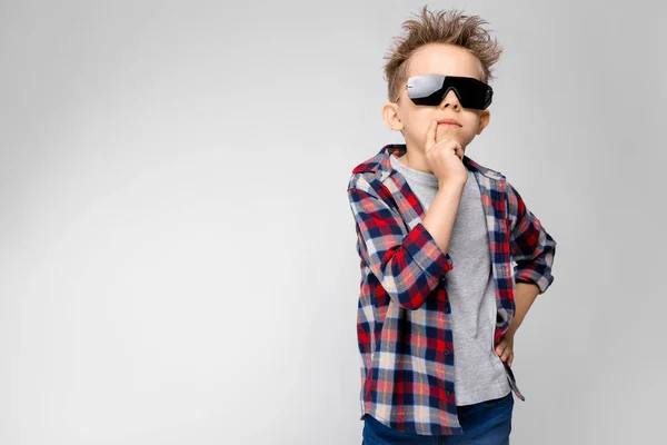 En stilig pojke i en rutig skjorta, grå skjorta och jeans står på en grå bakgrund. Pojken i svarta solglasögon. Pojken sätta fingret till munnen — Stockfoto