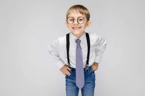 Ein charmanter Junge in weißem Hemd, Hosenträgern, Krawatte und heller Jeans steht vor grauem Hintergrund. der Junge mit der Brille lächelt — Stockfoto