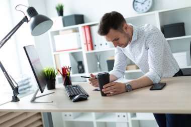 Genç bir adam bir bardak kahve ve bir kalem tutan bir tablo ofiste, yakın duruyor. Genç bir adam belgeleri ve bir bilgisayar ile çalışır.