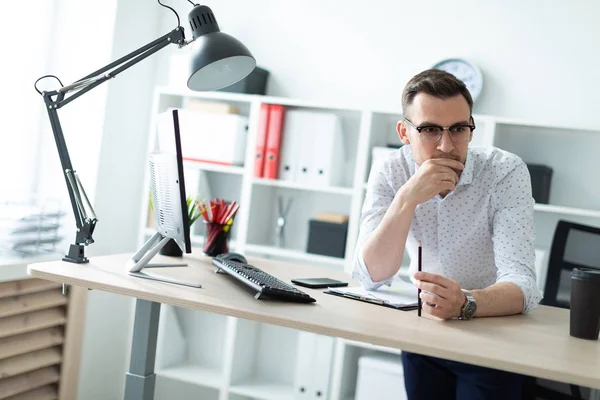 Un joven con gafas se para cerca de una mesa en la oficina y lleva un lápiz en la mano. . — Foto de Stock