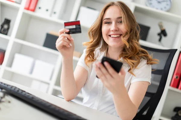 Hermosa joven se sienta en la oficina, tiene una tarjeta bancaria y teléfono en la mano . — Foto de Stock